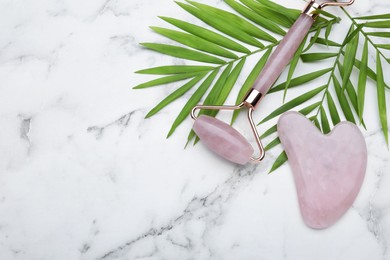 Photo of Gua sha stone, face roller and green leaves on white marble table, flat lay. Space for text