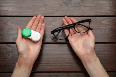 Photo of Woman holding case with contact lenses and glasses at wooden table, top view