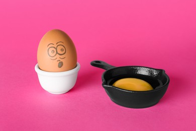 Photo of Egg with drawn shocked face and yolk in small frying pan on pink background