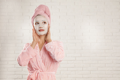 Young woman in bathrobe with cotton facial mask near white brick wall. Space for text