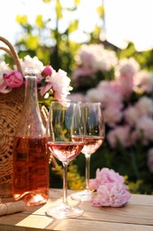 Bottle and glasses of rose wine near beautiful peonies on wooden table in garden