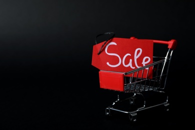 Tag with word Sale and toy shopping cart on dark background, space for text. Black friday concept