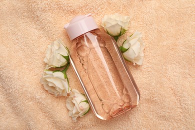 Photo of Bottle of micellar cleansing water and flowers on pink towel, flat lay