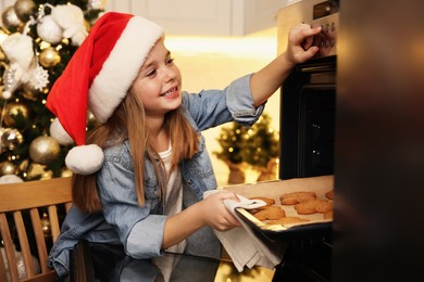 Little child in Santa hat taking baking sheet with Christmas cookies out of oven indoors