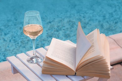Photo of Glass of tasty wine and open book on wooden table near swimming pool