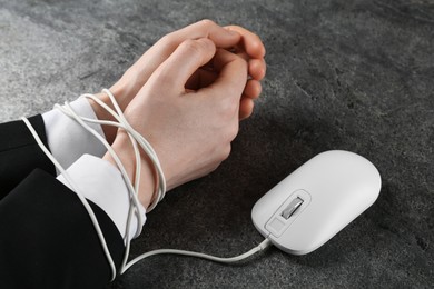 Man showing hands tied with computer mouse cable at grey table, closeup. internet addiction
