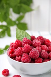 Photo of Bowl of fresh ripe raspberries with green leaf on white table against blurred background, closeup