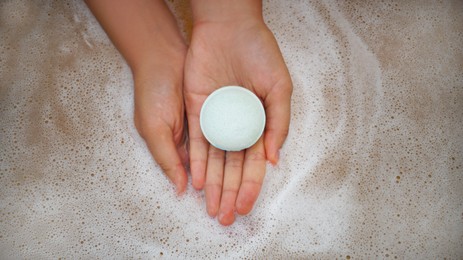 Woman holding bath bomb over water with foam, top view
