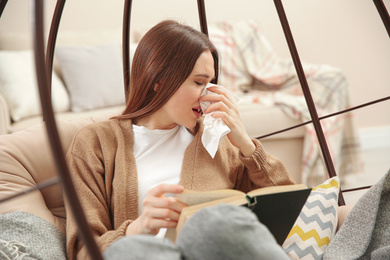 Sick young woman sneezing while reading book at home. Influenza virus