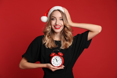 Happy young woman wearing Santa hat with alarm clock on red background. Christmas time