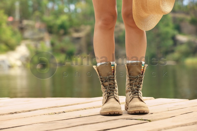 Young woman with hat on wooden pier near lake, focus on legs. Camping season