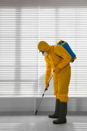 Male worker in protective suit spraying insecticide on wooden floor indoors. Pest control