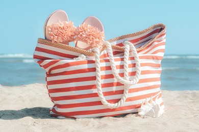 Photo of Stylish striped bag with slippers and seashell on sandy beach near sea