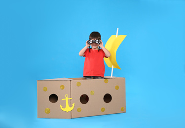 Cute little child playing with binoculars and cardboard ship on light blue background