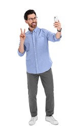 Photo of Smiling man taking selfie with smartphone and showing peace sign on white background