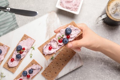 Woman holding tasty cracker sandwich with cream cheese, blueberries, red currants and thyme at grey table, top view