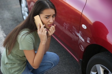 Stressed woman talking on phone near car with scratch outdoors