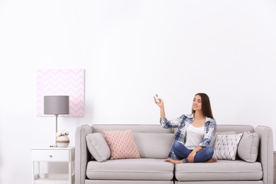 Photo of Young woman switching on air conditioner while sitting on sofa near white wall