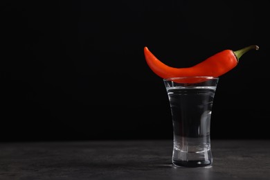 Red hot chili pepper and vodka in shot glass on grey table against black background, space for text