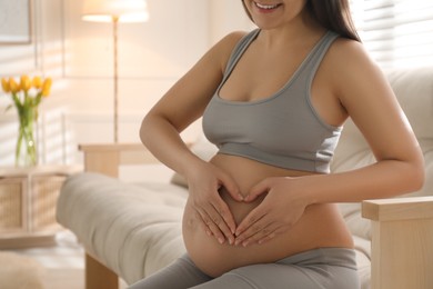 Pregnant young woman making heart with her hands on belly at home, closeup. Space for text