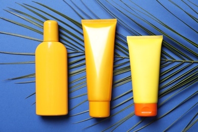 Photo of Different sun protection cosmetic products and green palm leaf on blue background, flat lay