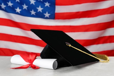 Graduation hat and diploma on light grey table against flag of United States