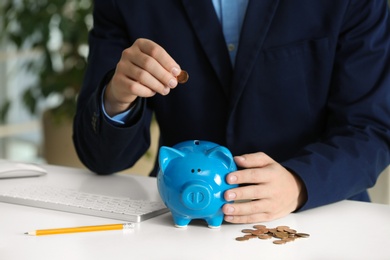 Man putting coin into piggy bank at white table, closeup. Money savings