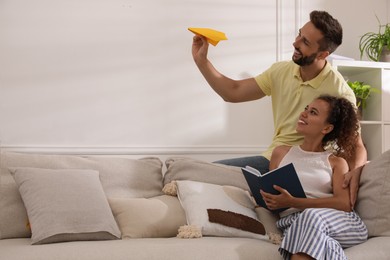 Happy man playing with paper plane while his girlfriend reading book on sofa in living room