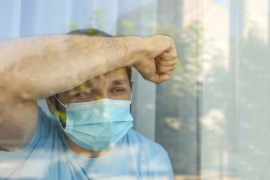 Stressed man in protective mask looking out of window, view through glass. Self-isolation during COVID-19 pandemic