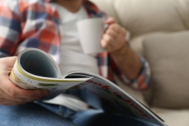 Man with cup of drink reading magazine on sofa, closeup