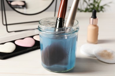 Cleaning makeup brushes in jar with special liquid on white wooden table, closeup