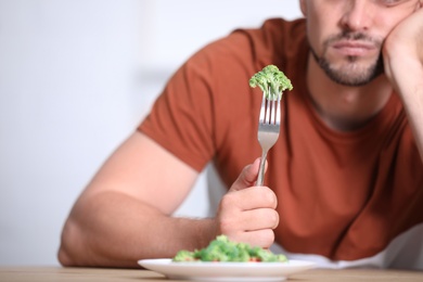 Unhappy man with broccoli on fork at table, closeup