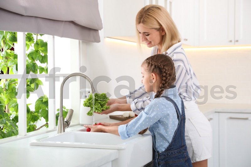 Little girl with her mother washing vegetables together in modern kitchen