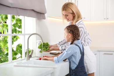 Little girl with her mother washing vegetables together in modern kitchen