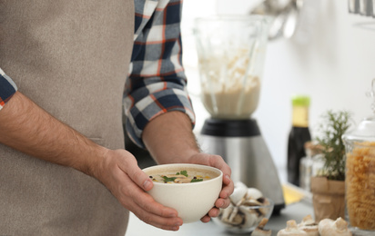 Young man holding bowl of tasty cream soup in kitchen, closeup