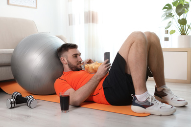 Lazy young man with smartphone eating junk food on yoga mat at home