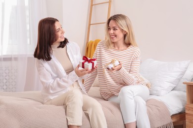 Photo of Smiling young women presenting gifts to each other on bed at home