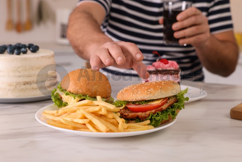 Overweight man with glass of cold drink taking burger from plate at table in kitchen, closeup