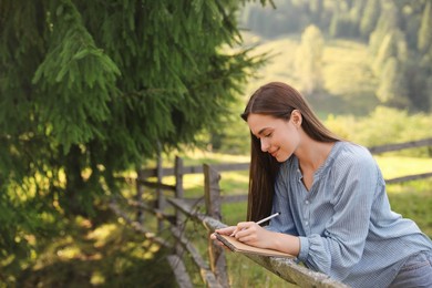 Beautiful young woman drawing with pencil in notepad near wooden fence