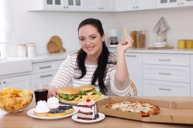 Happy overweight woman with unhealthy food in kitchen