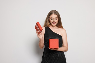 Emotional young woman in elegant black dress opening red gift box on white background