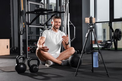 Photo of Man with ball and dumbbells streaming online training on phone at gym. Fitness coach