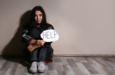 Abused young woman with sign HELP near beige wall. Domestic violence concept