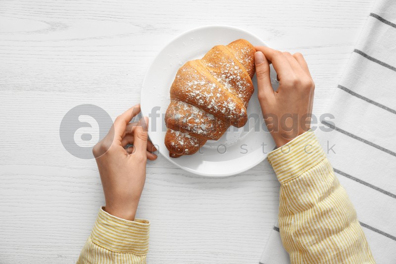 Woman eating fresh croissant at white wooden table, top view. Delicious morning meal