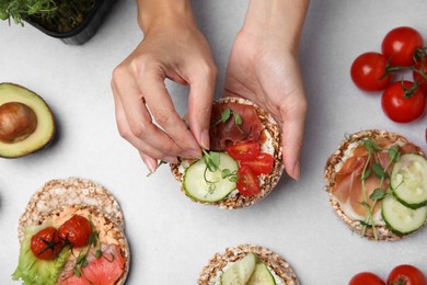 Woman adding microgreens onto crunchy buckwheat cakes with prosciutto, pieces of tomato and cucumber slice at white table, closeup