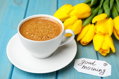 Cup of aromatic coffee, beautiful yellow tulips and Good Morning note on light blue wooden table