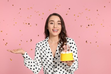 Photo of Coming of age party - 18th birthday. Smiling woman holding delicious cake with number shaped candles and catching confetti on pink background