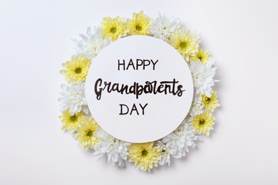 Card with phrase Happy Grandparents Day and beautiful flowers on white background, flat lay