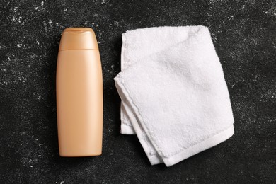 Soft folded terry towel and cosmetic bottle on black textured background, flat lay