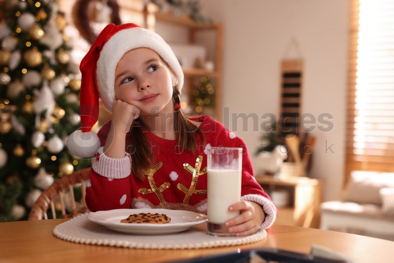 Cute little girl in Santa hat with delicious Christmas cookie and milk at home, space for text